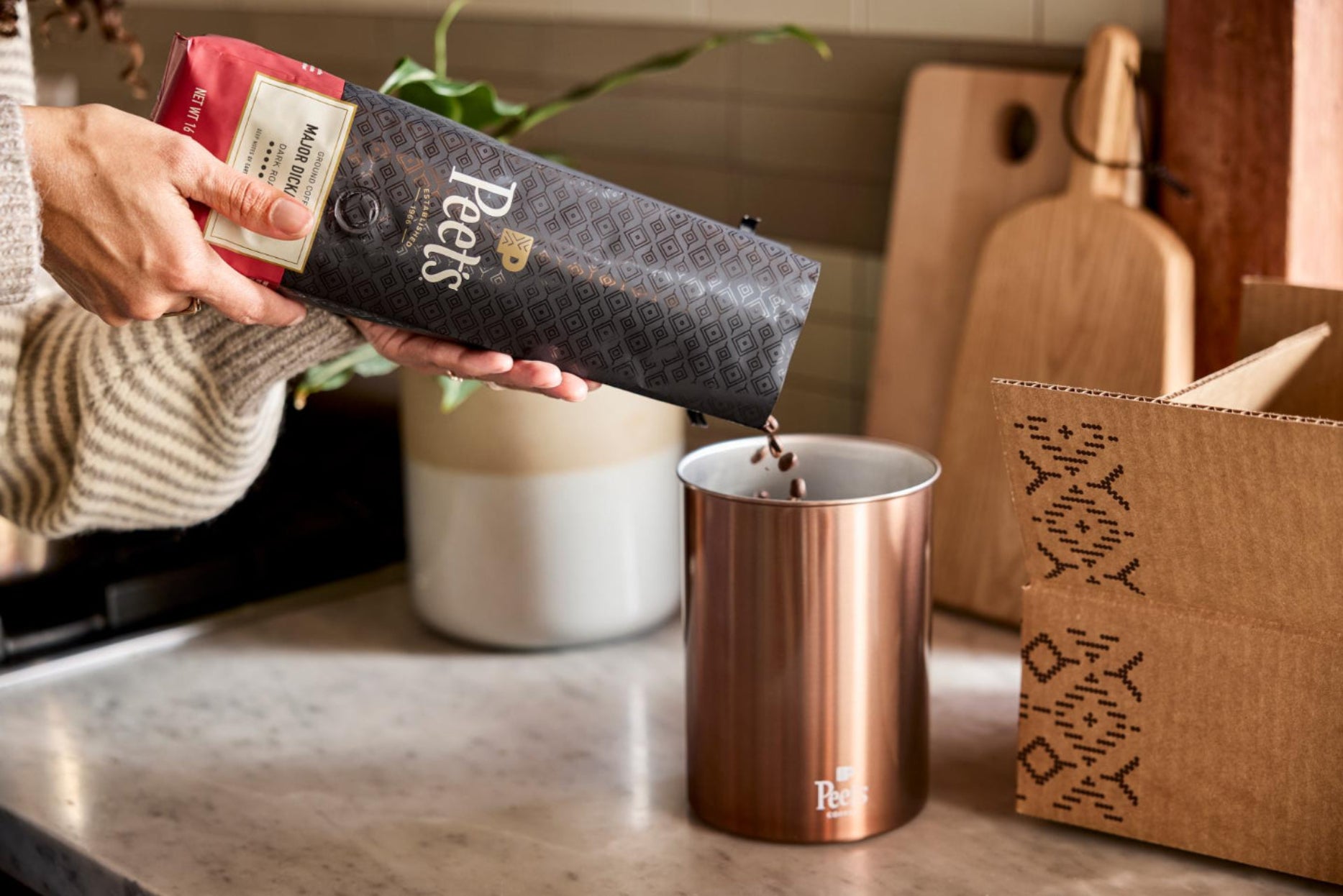 A person's hands pour coffee from a Peets paper pouch into a copper-colored metal cup on a kitchen counter, with a branded cardboard box nearby.